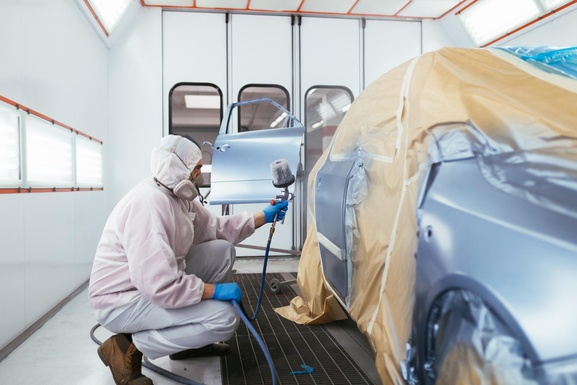 Ein Fahrzeuglackierer im Schutzanzug lackiert ein Auto in einer professionellen Lackierkabine. Das Auto ist teilweise mit Abdeckpapier geschützt, während die Tür und andere Bereiche in Metallic-Blau gespritzt werden
